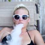 A woman in a tank top and sunglasses, reclining on an outdoor chair, exhales vapor from a CBD vape. Pure Relief Disposable CBD Vapes tasted great and left our reviewer feeling great too.