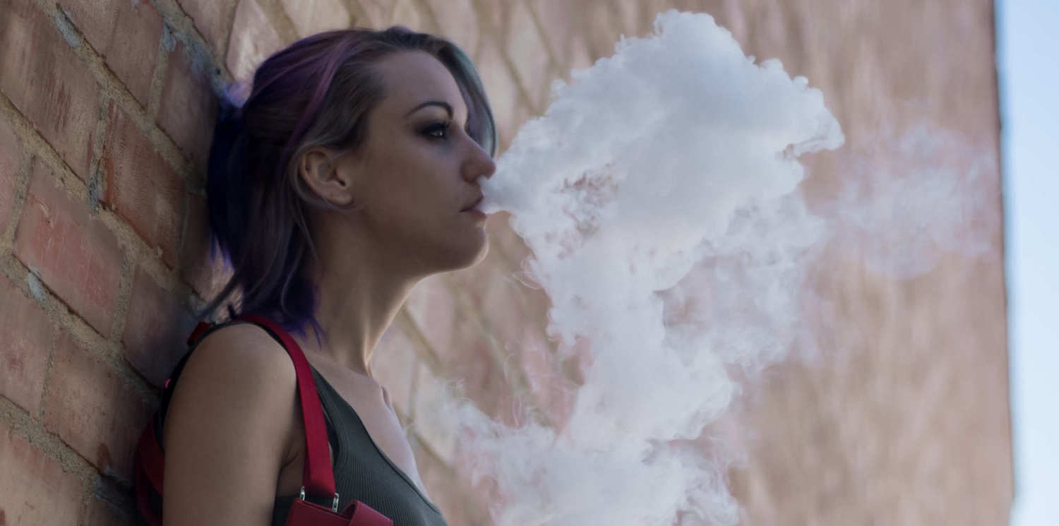 A woman leaning against a brick wall exhales after taking CBD vape oil. Vaping CBD is a fast, convenient way to take CBD oil.