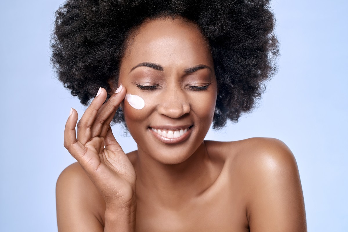 A black woman smiles with her eyes closed as she applies moisturizer to her cheek. More people are using hemp oil in skincare because of its numerous benefits.