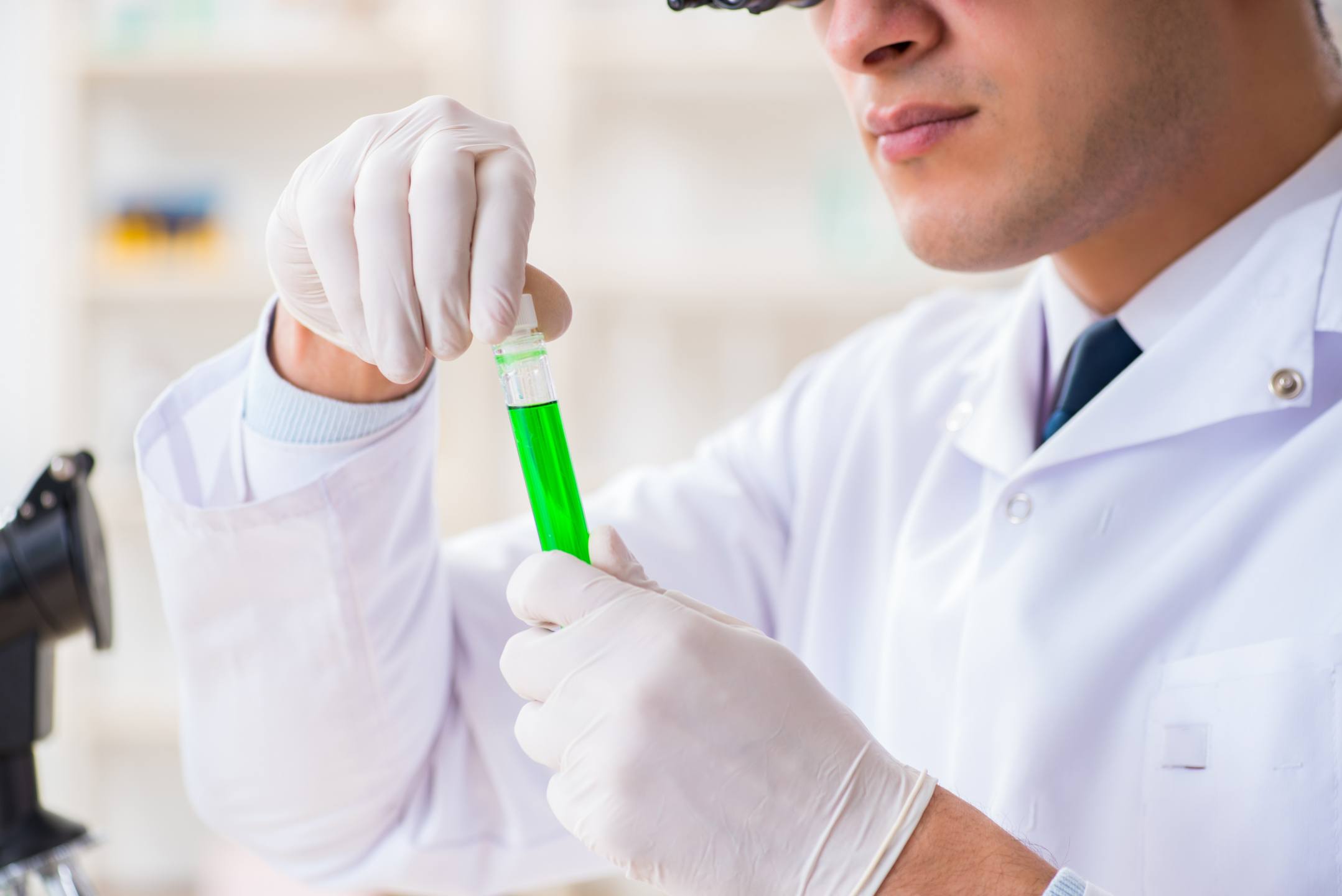 Hemp and cannabis testing labs are vital to building consumer trust, but results can vary dramatically from lab to lab. Photo: A scientist in gloves and a white lab coat examines a test tube of green liquid, with a microscope nearby.