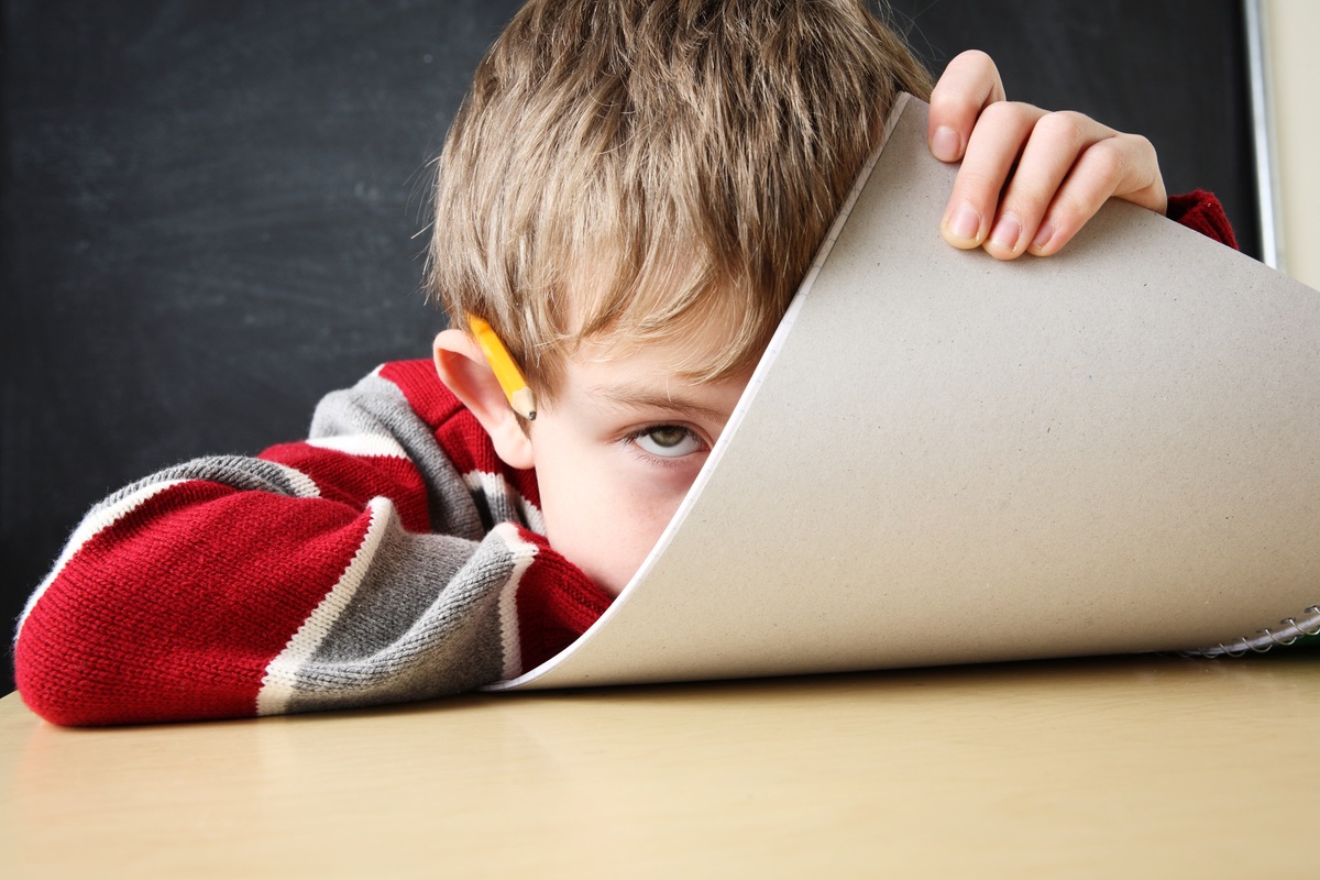 A young student hides playfully behind a piece of posterboard while sitting at a desk. Some research supports using cannabinoids like CBD for ADHD.