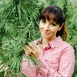 On the Ministry of Hemp Podcast, we're beginning a series on the women of hemp. Photo: A smiling young woman stands in a hemp field, wearing a pink button down. One hand holds part of a leafy hemp plant.