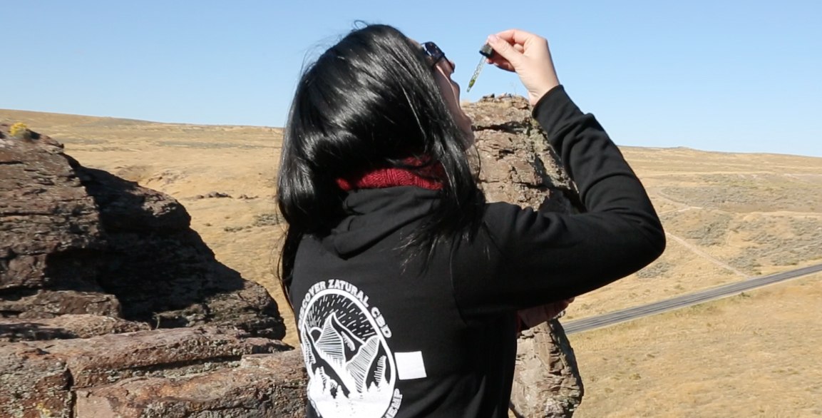 We were surprised by how effectively Zatural CBD Oil Drops helped us feel better. Photo: In a desert setting, a woman in a Zatural-branded hoodie takes CBD oil from a dropper.