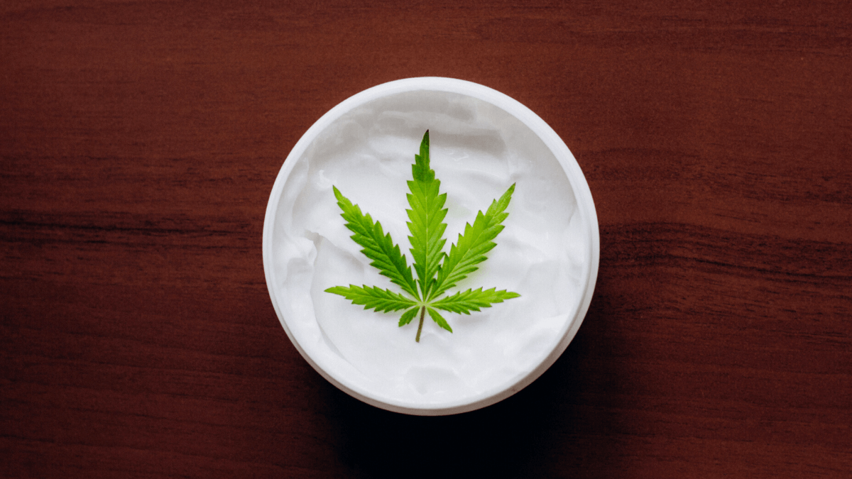 CBD topicals quickly became one of the most popular forms of CBD. Here's a closer look at why. Photo: A plastic container of a lotion CBD topical with a hemp leaf on it, sitting on a dark wooden table.