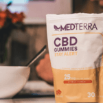 Medterra Stay Alert CBD Gummies on a desktop. In the background is a cereal bowl, soe flowers, and a person working on a desktop.