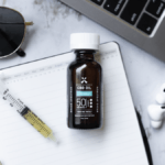 Green Roads Full Spectrum CBD Oil with measuring syringe sits on a desktop with soome earbuds, sunglasses, a notepad, and part of a laptop keyboard is also visible.